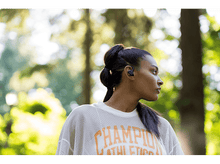 Charger l&#39;image dans la galerie, Femme avec des écouteurs regardant vers la droite, dans un parc, avec des arbres verts et du soleil.
