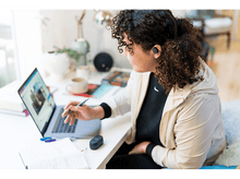 Charger l&#39;image dans la galerie, Une femme à un bureau avec un ordinateur portable et un stylo, portant des écouteurs, travaillant à domicile.
