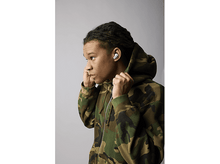 Charger l&#39;image dans la galerie, Un jeune homme portant un sweat à capuche camouflage met des écouteurs blancs, regardant vers la gauche sur fond gris.
