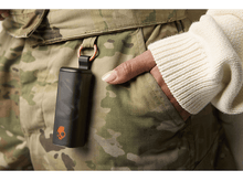 Charger l&#39;image dans la galerie, Un étui Skullcandy noir est suspendu à un pantalon camouflage. Une main repose dans une poche, une manche crème est visible.
