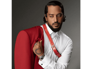 Un homme en chemise blanche et veste rouge, regardant de côté, avec un tatouage sur la main.