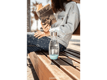 Charger l&#39;image dans la galerie, Personne lit un livre assise sur un banc. Bouteille d&#39;eau devant. Bulles flottent dans l&#39;air.
