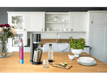 Charger l&#39;image dans la galerie, Scène de cuisine avec un soda maker, une bouteille, un verre et des tranches de lime sur un comptoir en bois.
