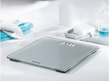 Charger l&#39;image dans la galerie, Pèse-personne en verre avec affichage numérique indiquant le poids. Fond blanc avec serviette et savon.
