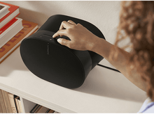 Charger l'image dans la galerie, Main d'une personne réglant le volume sur un haut-parleur Sonos noir sur une étagère avec des livres.
