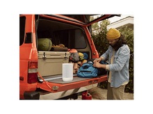 Charger l&#39;image dans la galerie, Un homme range un sac bleu dans un coffre de voiture. Il y a un haut-parleur et du matériel de camping.
