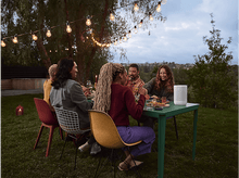 Charger l&#39;image dans la galerie, Un groupe de personnes est assis autour d&#39;une table, éclairé par des guirlandes lumineuses en extérieur.
