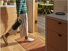 Charger l&#39;image dans la galerie, Personne en pull vert et pantalon blanc traversant une porte, tenant un cylindre noir.
