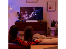 Charger l&#39;image dans la galerie, Scène de salon. TV sur un meuble, barre de son en dessous. Deux personnes sur un canapé regardent la TV.
