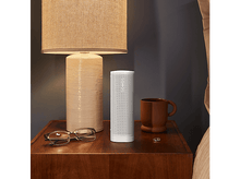 Charger l&#39;image dans la galerie, Haut-parleur blanc sur une table de chevet à côté d&#39;une lampe, de lunettes et d&#39;une tasse à café. Le fond est un lit.
