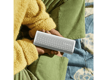 Charger l&#39;image dans la galerie, Une personne tient une enceinte Sonos blanche. Elle porte un pull jaune et un pantalon en velours vert.
