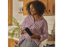 Charger l&#39;image dans la galerie, Femme tenant un haut-parleur noir près d&#39;une cuisine. Elle sourit et porte une chemise rayée violette.
