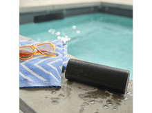 Charger l&#39;image dans la galerie, Haut-parleur noir sur le bord d&#39;une piscine avec lunettes et serviette, piscine en arrière-plan.
