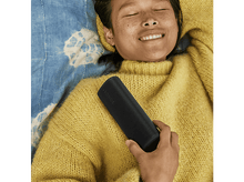 Charger l&#39;image dans la galerie, Personne allongée avec un haut-parleur noir. Elle porte un pull jaune et sourit. Couverture bleue en arrière-plan.
