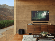 Charger l&#39;image dans la galerie, Salon avec une télévision sur un meuble en bois et une barre de son. Un caisson de basses noir au sol.
