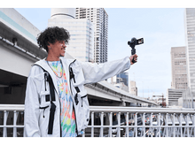 Charger l&#39;image dans la galerie, Homme tenant une caméra, souriant devant des bâtiments. Il porte une veste blanche et une chemise colorée.
