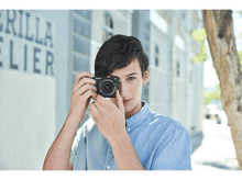 Charger l&#39;image dans la galerie, Un jeune homme tenant un appareil photo Sony noir, regardant à travers le viseur. Il est à l&#39;extérieur.
