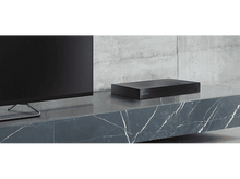 Charger l&#39;image dans la galerie, Un appareil Sony noir sur une surface en marbre, à côté d&#39;un écran de télévision. La pièce est minimaliste.
