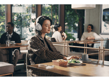 Charger l&#39;image dans la galerie, Une femme portant un casque blanc est assise à une table dans un café. Elle regarde vers le bas et travaille sur un ordinateur portable.
