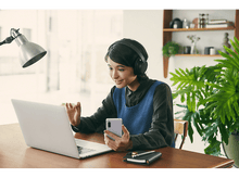 Charger l&#39;image dans la galerie, Femme en appel vidéo. Elle tient un téléphone et regarde un ordinateur portable sur un bureau en bois.
