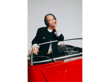 Charger l&#39;image dans la galerie, Une femme en veste noire et casque audio s&#39;appuie sur une voiture rouge. Elle semble pensive. Le fond est un mur blanc.
