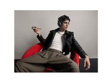 Charger l&#39;image dans la galerie, Un homme en veste en cuir et casque est assis dans une chaise rouge, tenant un téléphone. Il porte des bagues et une chaîne. Fond gris.
