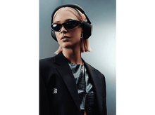 Charger l&#39;image dans la galerie, Une femme portant des lunettes de soleil, un casque, un blazer noir et un haut à motif reptile, avec les cheveux blonds, sur fond gris.
