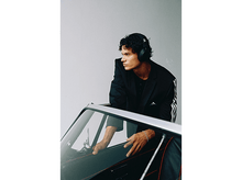 Charger l&#39;image dans la galerie, Un homme en veste noire et casque audio s&#39;appuie sur une voiture. Il a les cheveux bouclés et un bracelet argenté. Logo Adidas visible.
