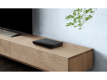 Charger l&#39;image dans la galerie, Un lecteur multimédia noir est posé sur un meuble en bois. Un téléviseur est visible à gauche, une plante à droite.
