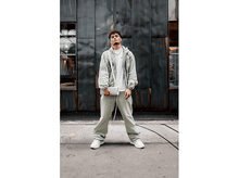 Charger l&#39;image dans la galerie, Un homme en vêtements vert clair debout devant un mur en métal gris texturé, tenant un petit sac blanc.
