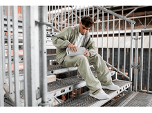 Charger l&#39;image dans la galerie, Un jeune homme assis sur des escaliers, tenant un haut-parleur blanc, portant une veste verte.
