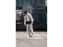 Charger l&#39;image dans la galerie, Un jeune homme en tenue décontractée se tient devant un mur industriel bleu, tenant un sac en bandoulière noir.
