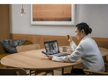 Charger l&#39;image dans la galerie, Femme en appel vidéo sur un ordinateur portable, tenant un téléphone, assise à une table en bois.
