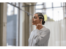 Charger l&#39;image dans la galerie, Femme aux cheveux tressés portant des écouteurs, se touchant l&#39;oreille avec son doigt. Dans un cadre de type bureau.
