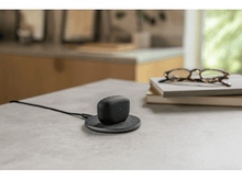 Charger l&#39;image dans la galerie, Écouteurs en charge sur un socle de chargement sans fil. Lunettes sur une pile de livres.
