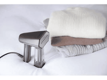 Charger l&#39;image dans la galerie, Vapeur à main sur un lit avec des pulls pliés. La vapeur est argentée et noire. Les pulls sont blancs, marrons et gris.
