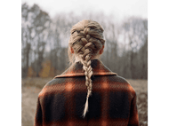 Dos d'une femme, avec une longue tresse blonde. Vêtue d'une veste à carreaux, debout dans un champ flou.