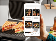 Charger l&#39;image dans la galerie, Une main tenant un téléphone affichant des recettes avec un grill en arrière-plan.
