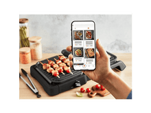 Charger l&#39;image dans la galerie, Une personne tenant un téléphone montrant des recettes de cuisine à côté d&#39;un grill avec des brochettes.
