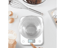 Charger l'image dans la galerie, Une balance de cuisine avec un bol en verre et une main appuyant sur un bouton. Ingrédients et ustensiles de cuisine.
