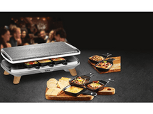 Charger l&#39;image dans la galerie, Grill à raclette avec aliments. Personnes floues en arrière-plan. Fromage, légumes et ingrédients visibles.
