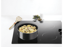 Charger l&#39;image dans la galerie, Pâtes dans une casserole sur une plaque à induction noire. D&#39;autres ingrédients et une cuillère en bois sont à proximité.
