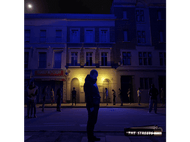 Scène de nuit avec des bâtiments et des gens. Un homme au premier plan. Panneau 'The Streets'.