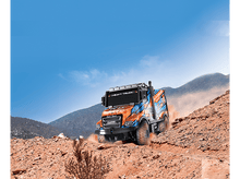 Charger l&#39;image dans la galerie, Un camion jouet rouge et orange roulant sur un chemin de terre, montagnes et ciel en arrière-plan.
