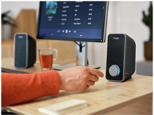 Charger l&#39;image dans la galerie, Une personne ajuste le volume d&#39;un haut-parleur Trust noir. Un écran d&#39;ordinateur est en arrière-plan.
