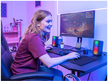 Charger l'image dans la galerie, Jeune femme jouant avec un moniteur, des haut-parleurs, un clavier et une souris. Éclairage violet.
