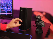 Charger l&#39;image dans la galerie, Une personne tourne le bouton de volume d&#39;un haut-parleur noir. Un buste est sur le bureau. Fond rose et rouge.
