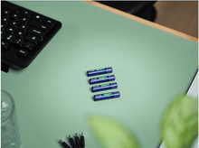 Charger l&#39;image dans la galerie, Quatre piles AA Trust sont sur un bureau vert clair. Un clavier, un verre et une plante sont sur le bureau.
