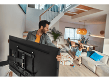 Charger l&#39;image dans la galerie, Homme portant une TV, enfant court. Intérieur d&#39;une maison avec escalier et meubles visible.
