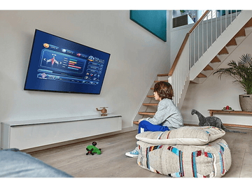 Un garçon regarde la télé au mur, un dinosaure et une voiture jouet sont par terre.